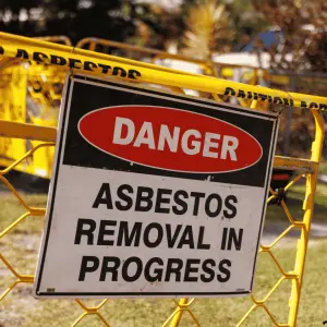 Warning sign reading 'DANGER: ASBESTOS REMOVAL IN PROGRESS' mounted on yellow safety fencing