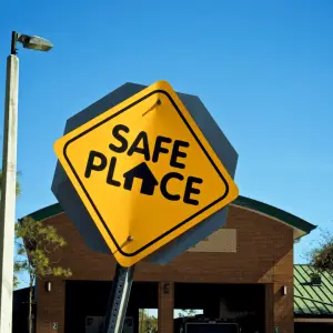 Let me provide the key text components for making this image accessible: Alt Text: "Yellow diamond-shaped road sign reading 'SAFE PLACE' with a house icon, mounted in front of a brick building on a clear day