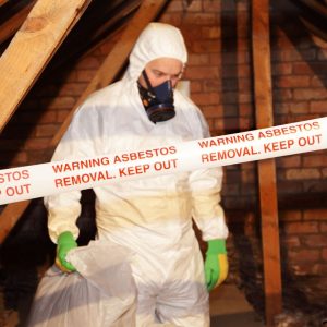 Professional in white protective suit and respirator performing asbestos removal in an attic, with red warning tape visible.